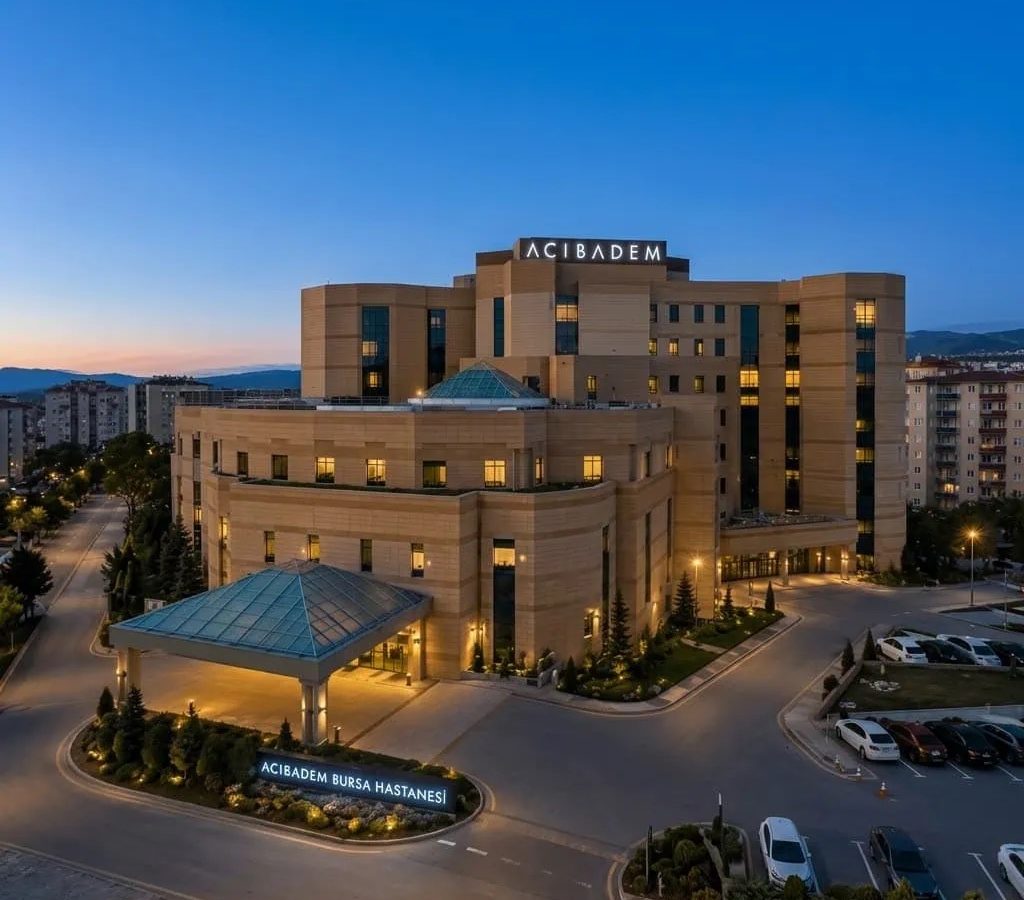 Exterior view of Acibadem Bursa Hospital at dusk with cityscape in background.